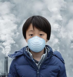 A child with a face mask standing in front of a row of chimneys with smoke rising from them
