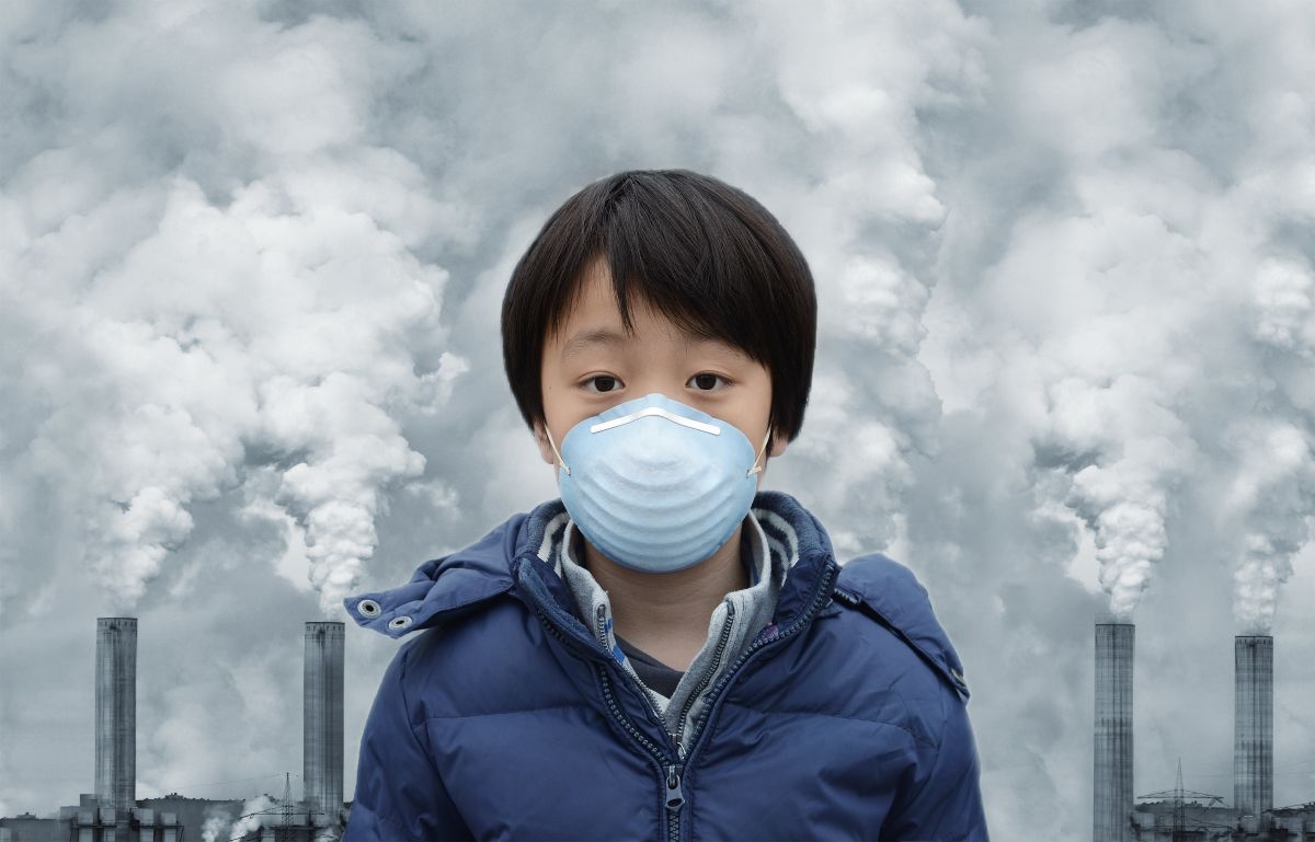 A child with a face mask standing in front of a row of chimneys with smoke rising from them